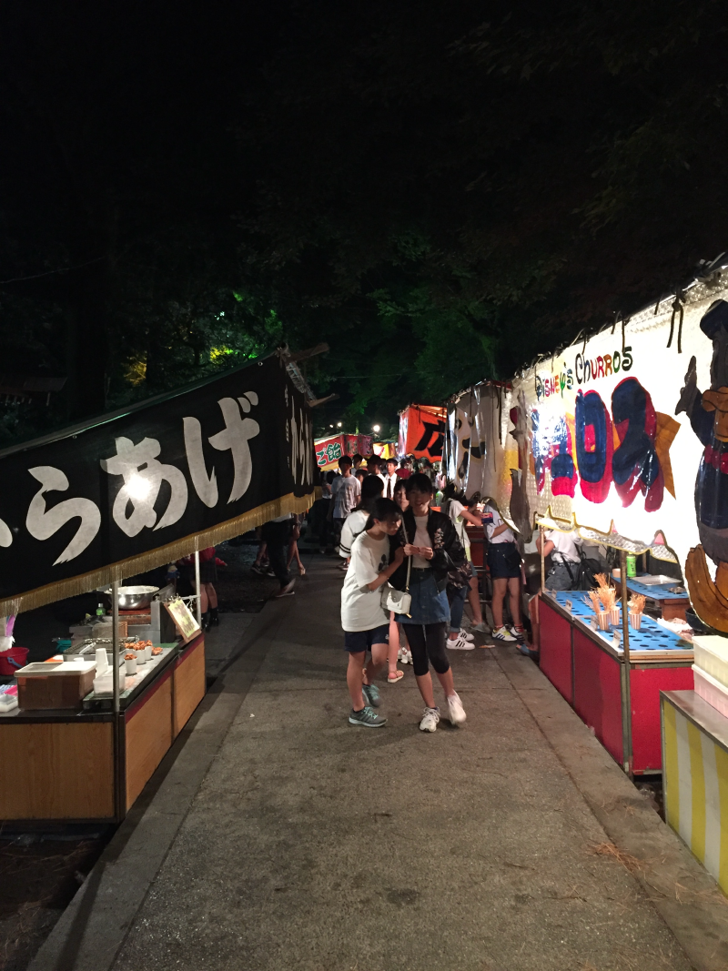 Two girls walk down a lane of lit festival stalls selling karaage and churros at night