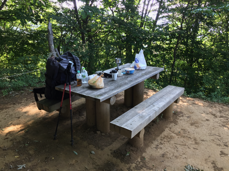 Breakfast laid out on a picnic table --- stove, food bag and drinks beside a backpack and trekking poles