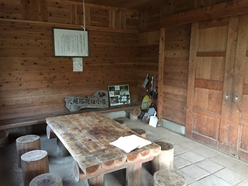 Cedar-walled hut interior with log stools around a heavy plank table and a carved 越路避難小屋 sign on the bench