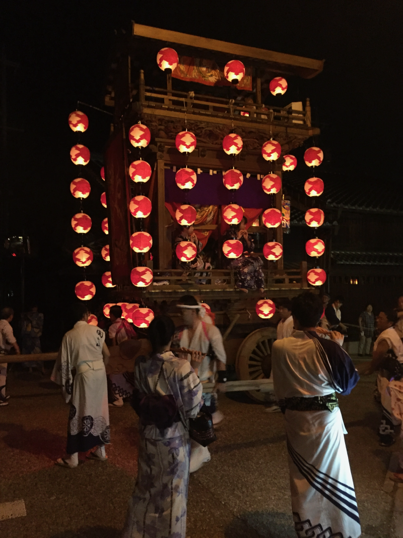 Towering festival float draped with rows of red paper lanterns, surrounded by people in yukata with a flute player in the foreground