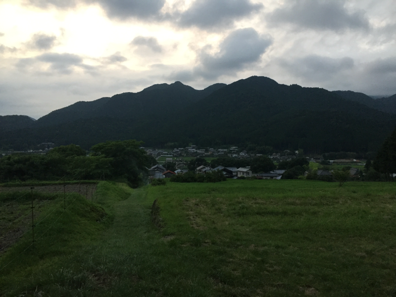 Small village nestled between grassy fields and dark wooded hills under a cloudy dusk sky