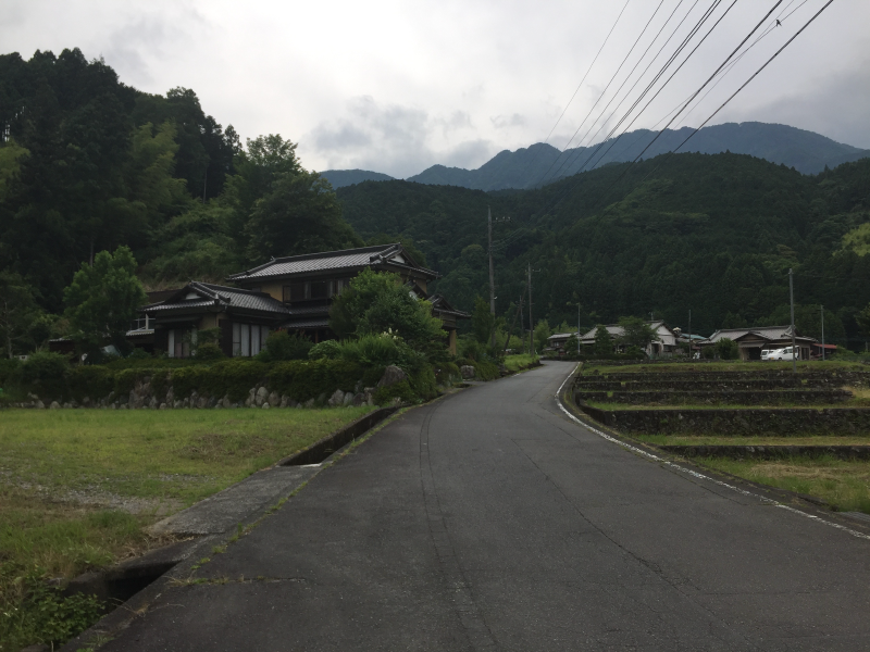 Paved lane curving past a traditional Japanese farmhouse, terraced rice paddies and forested mountains beyond